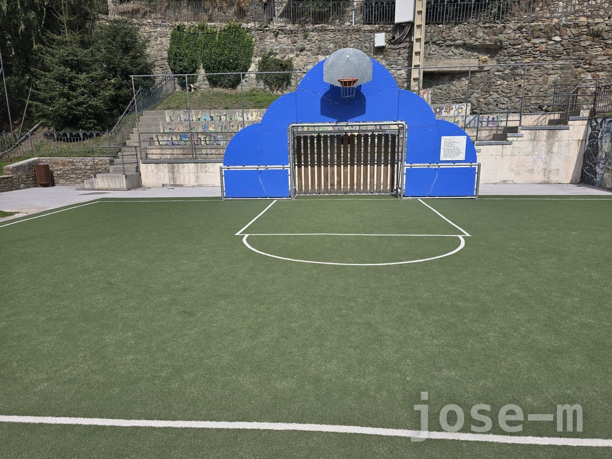 Pista de césped artificial verde en Callissa de Som, con una canasta sobre un panel azul que corona una portería de fútbol y un muro de piedra detrás