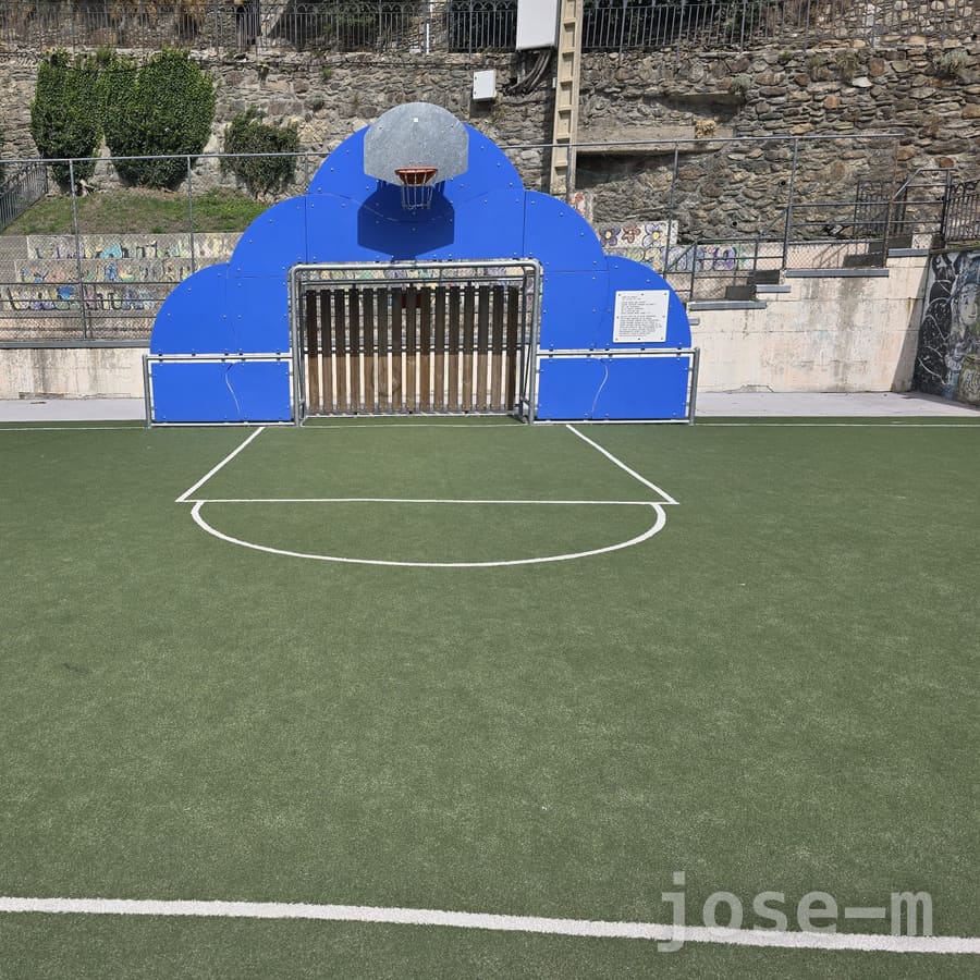 Pista de césped artificial verde en Callissa de Som, con una canasta sobre un panel azul que corona una portería de fútbol y un muro de piedra detrás