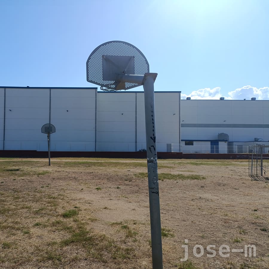 Canasta de tablero metálico perforado en primer plano y otra al fondo, sobre un descampado de hierba seca frente a una nave industrial blanca, en el Passeig Fluvial de Granollers