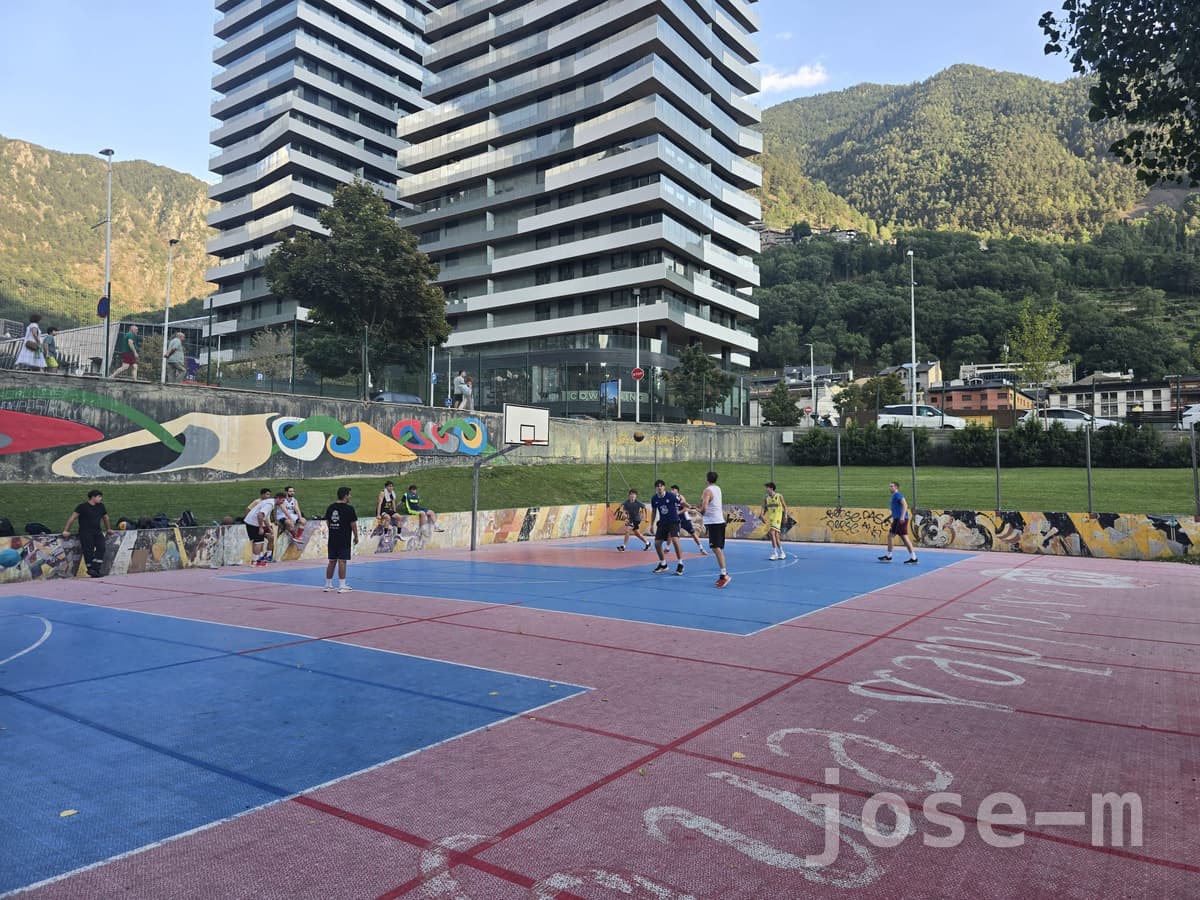 Cancha exterior de pavimento azul y rojo en Prat de Roure, con un partido en marcha, un mural de colores en el muro de contención y una torre de viviendas entre montañas al fondo