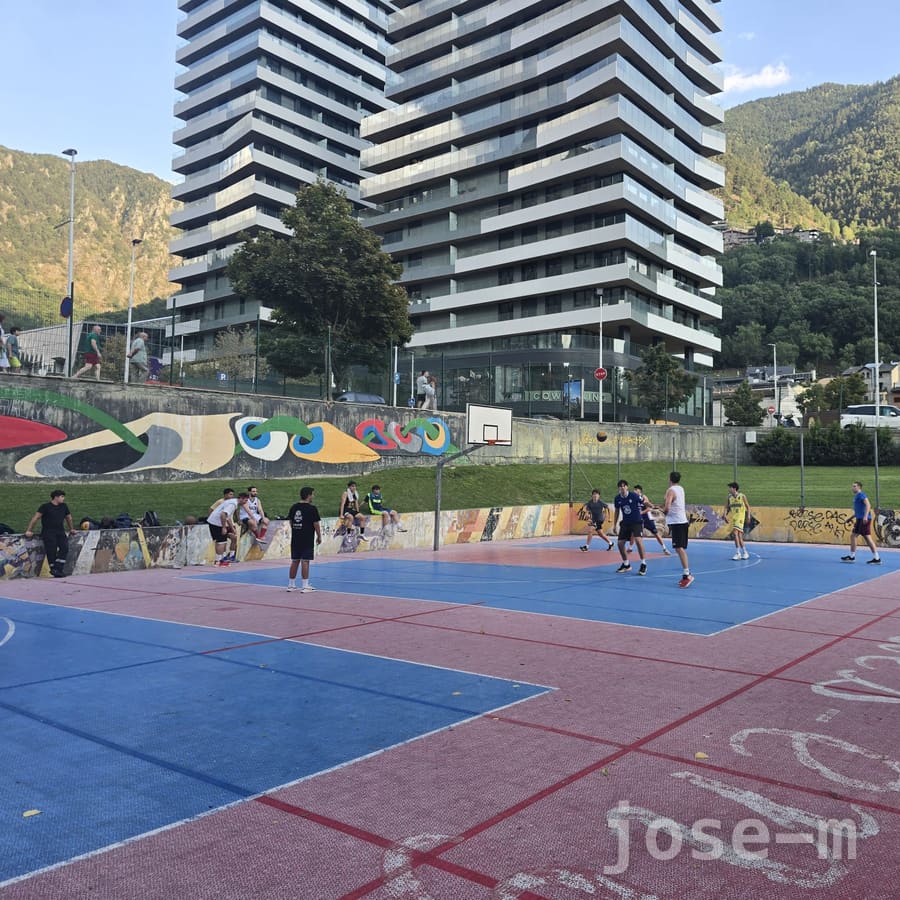 Cancha exterior de pavimento azul y rojo en Prat de Roure, con un partido en marcha, un mural de colores en el muro de contención y una torre de viviendas entre montañas al fondo